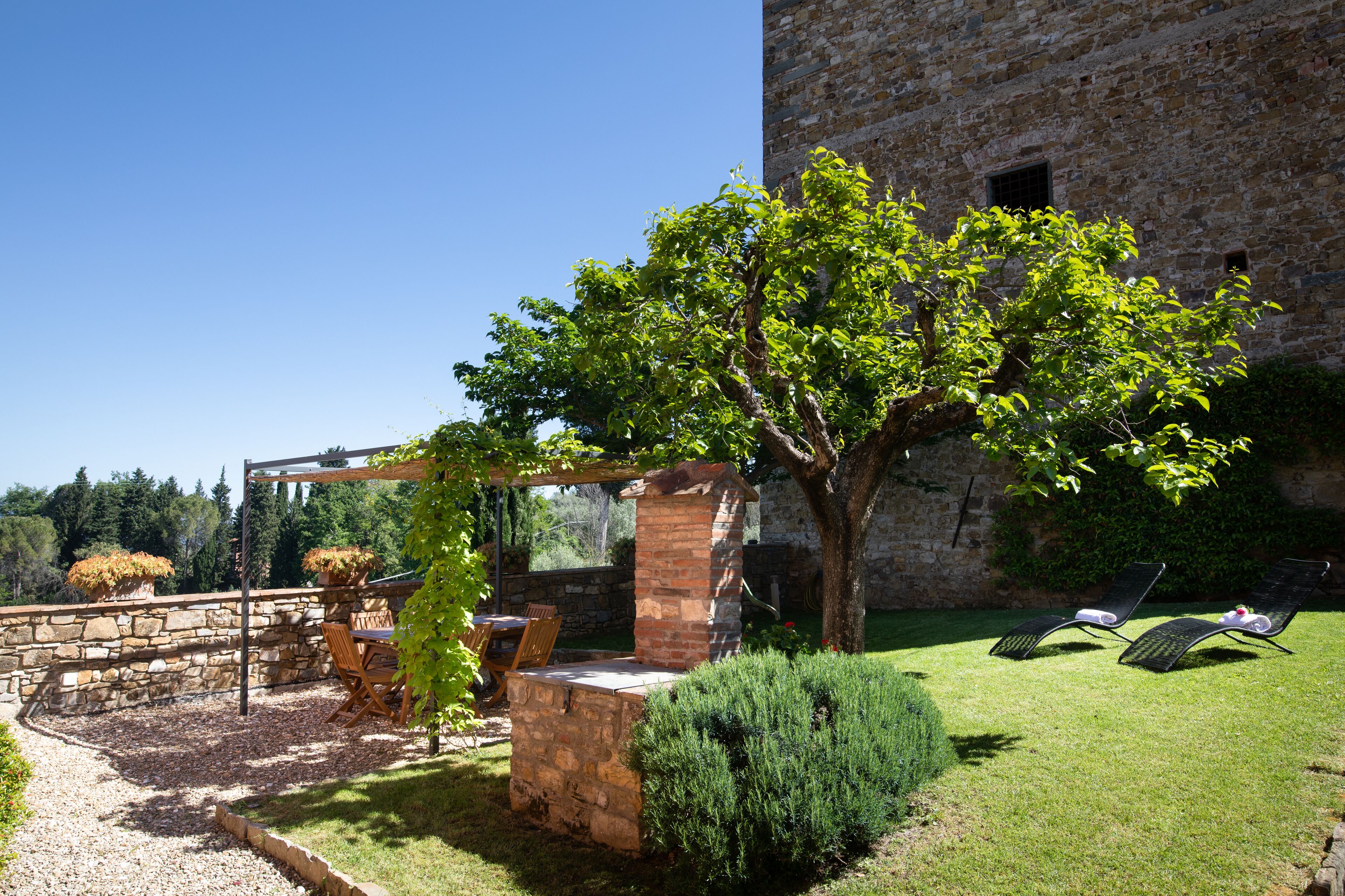 Un edificio in pietra con un'area barbecue in mattoni, un pergolato con sedie e un prato curato con due poltrone da relax.