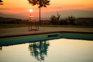 Una piscina con mesa y sillas, rodeada de un jardín ajardinado y una vista al atardecer.
