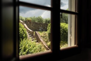 Vista a través de una ventana que muestra un muro de piedra y una escalera de piedra que sube a un nivel superior.
