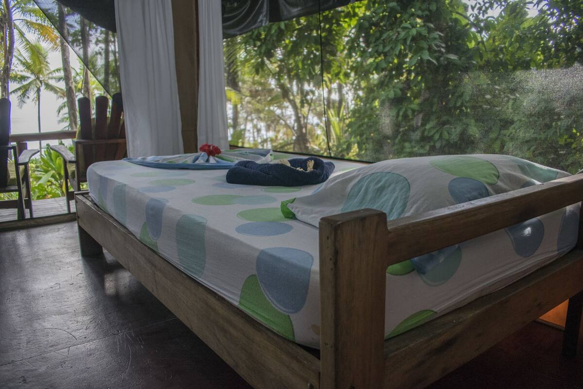 A wooden bed with a patterned bedspread, a small table with a red flower, and a view of lush greenery through a window.