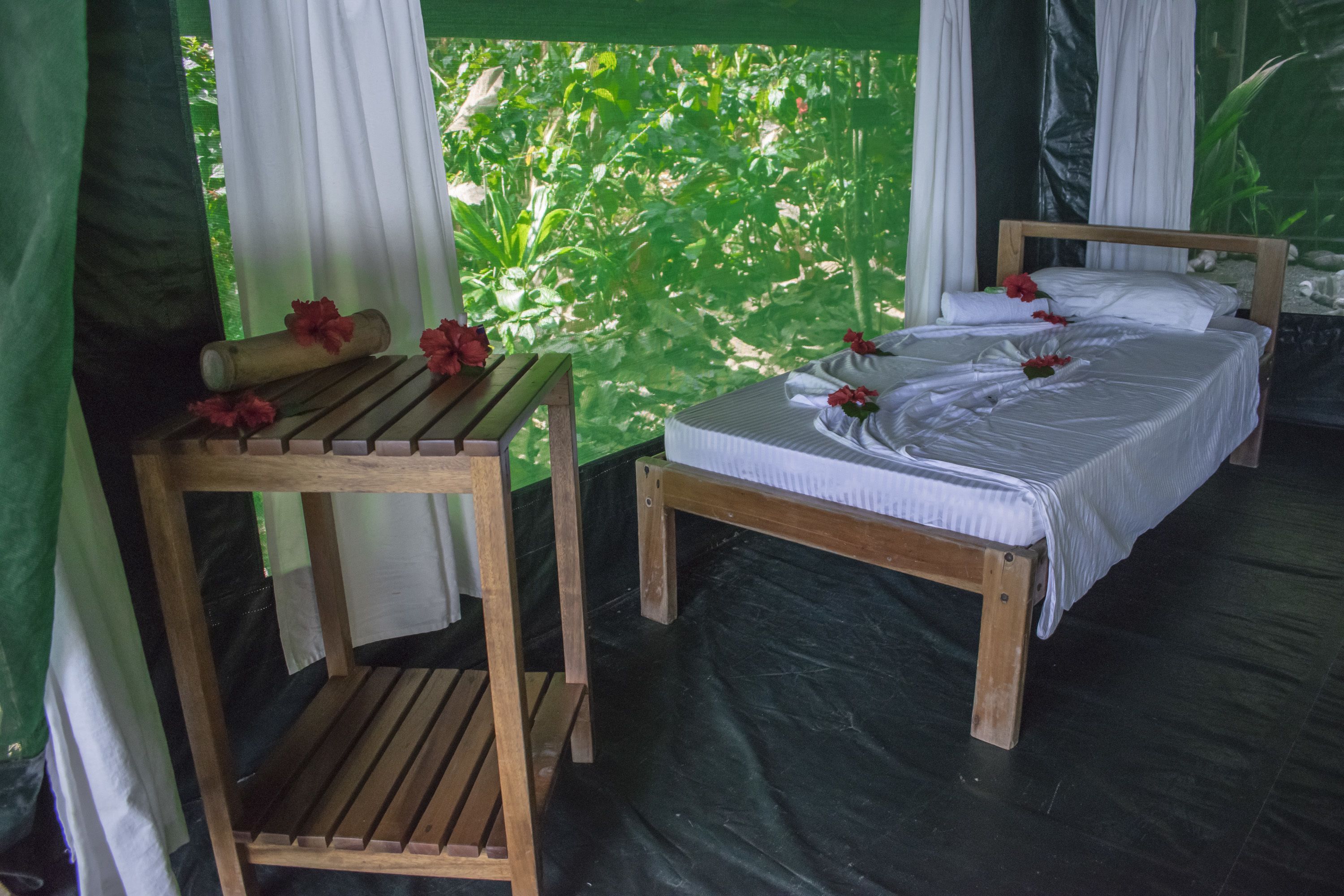 A single bed with white linens and a wooden headboard, a wooden table with a rolled-up mat and a red flower, and a view of lush greenery through a window with white curtains.