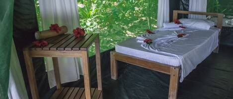 A single bed with white linens and a wooden headboard, a wooden table with a rolled-up mat and a red flower, and a view of lush greenery through a window with white curtains.