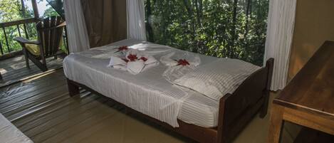 A neatly made bed with white linens and red flowers, a wooden chair, and a view of lush greenery through large windows.