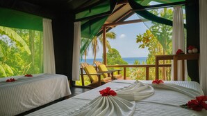 Een hotelkamer met twee bedden, een balkon met uitzicht op zee en tropische planten.