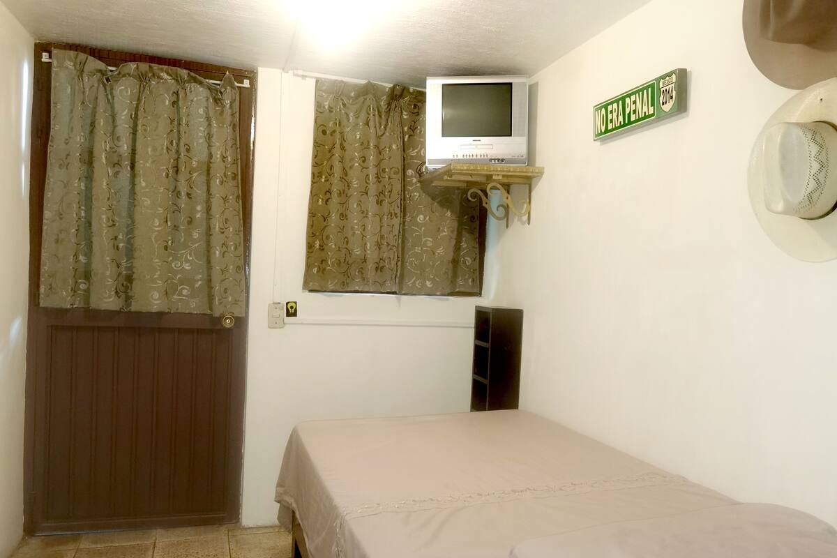 Una stanza piccola e modesta con un letto, una televisione montata su uno scaffale sopra una finestra e una porta con tende.