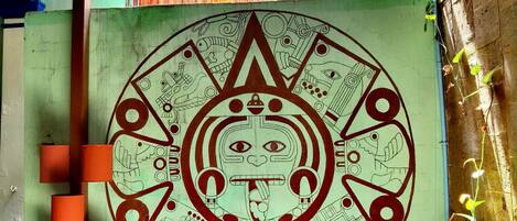 A room with a large mural featuring a circular design and an eagle. There are orange cylindrical objects on the left and a wooden structure at the bottom.