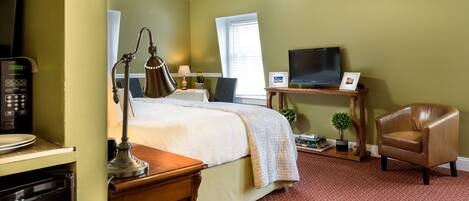 A hotel room with a bed, a desk, a chair, a television, and a small table with a plant and books.