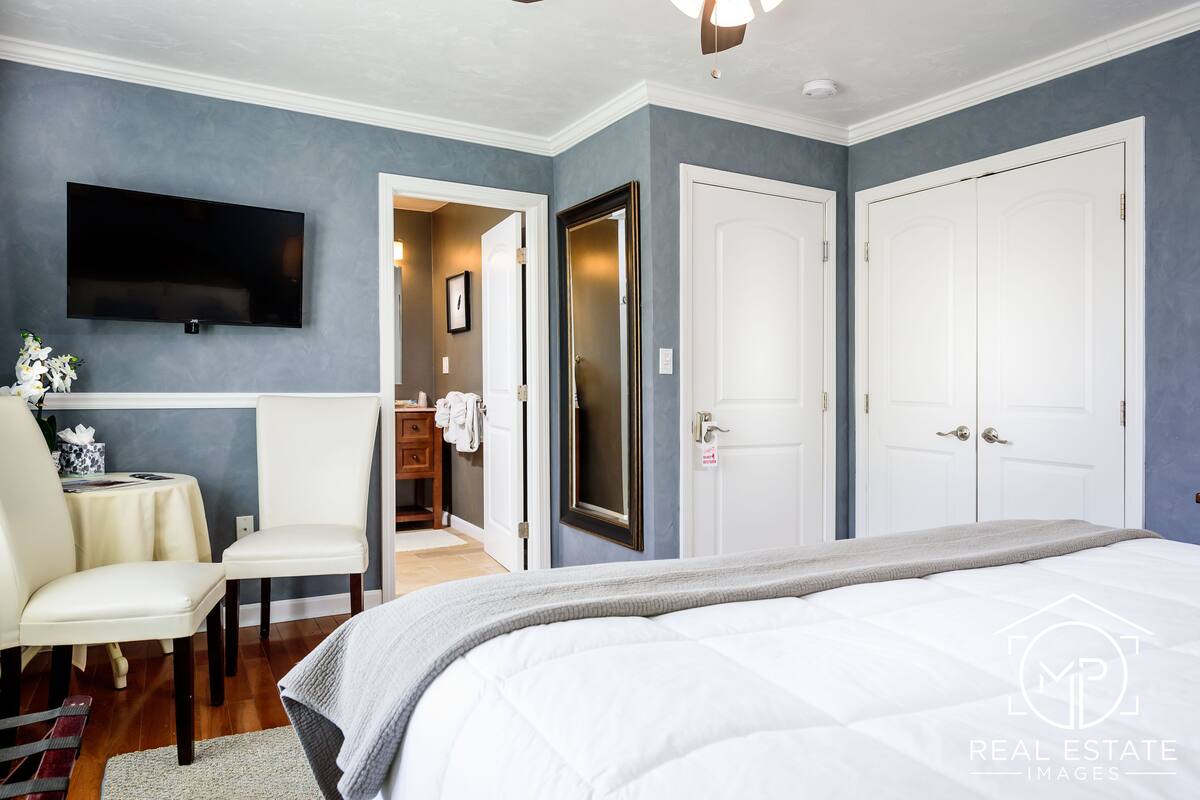 A bedroom with a bed, a TV mounted on the wall, a chair, and a small table with a vase of flowers.