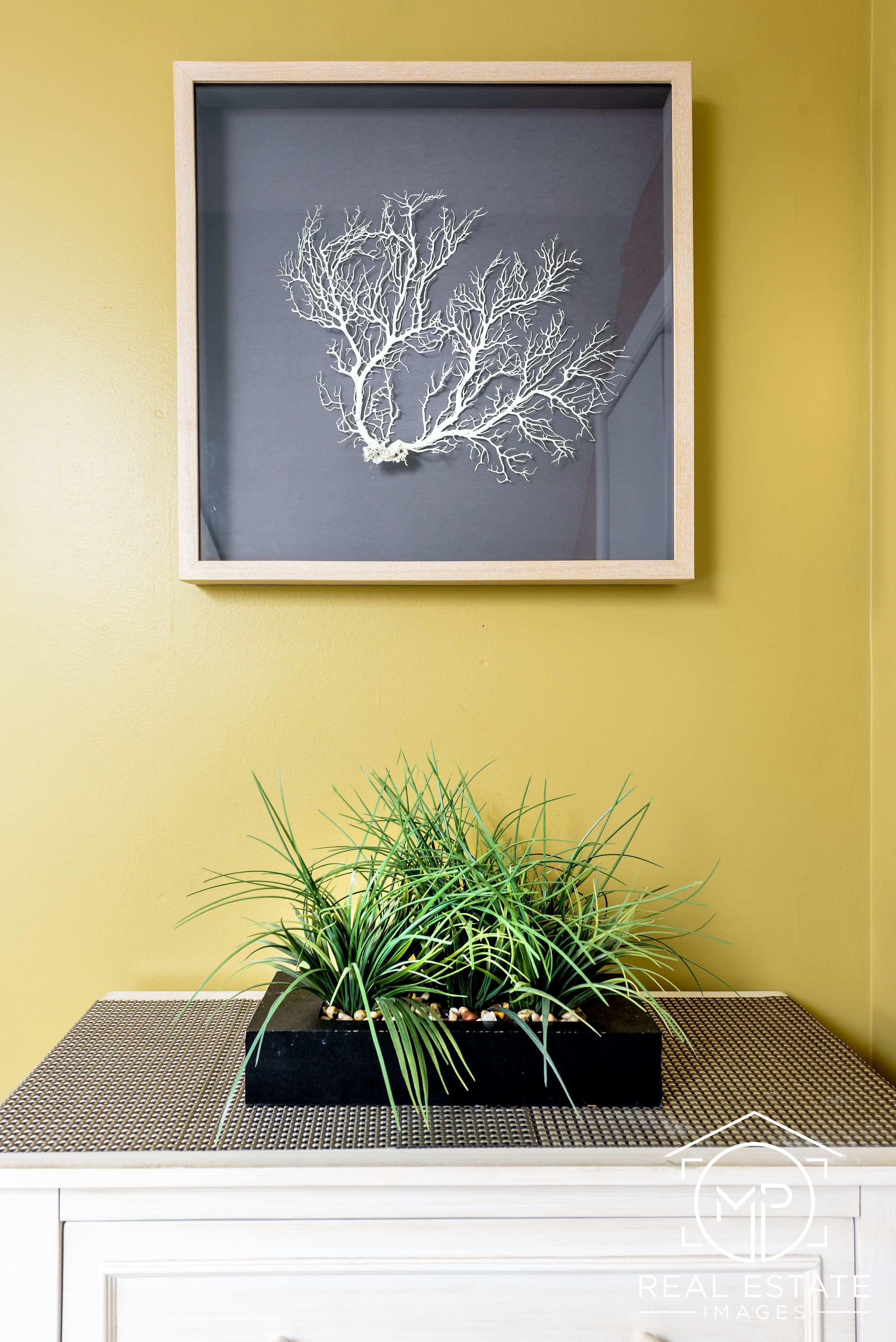 Una obra de arte enmarco con un árbol en la pared, una planta en maceta sobre una mesa y un aparador blanco.