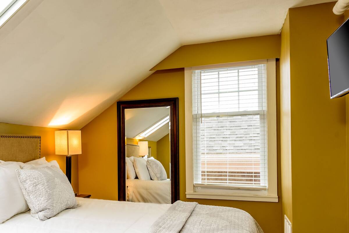 A bedroom with a skylight, a bed with pillows, a bedside lamp, a mirror, and a window with blinds.