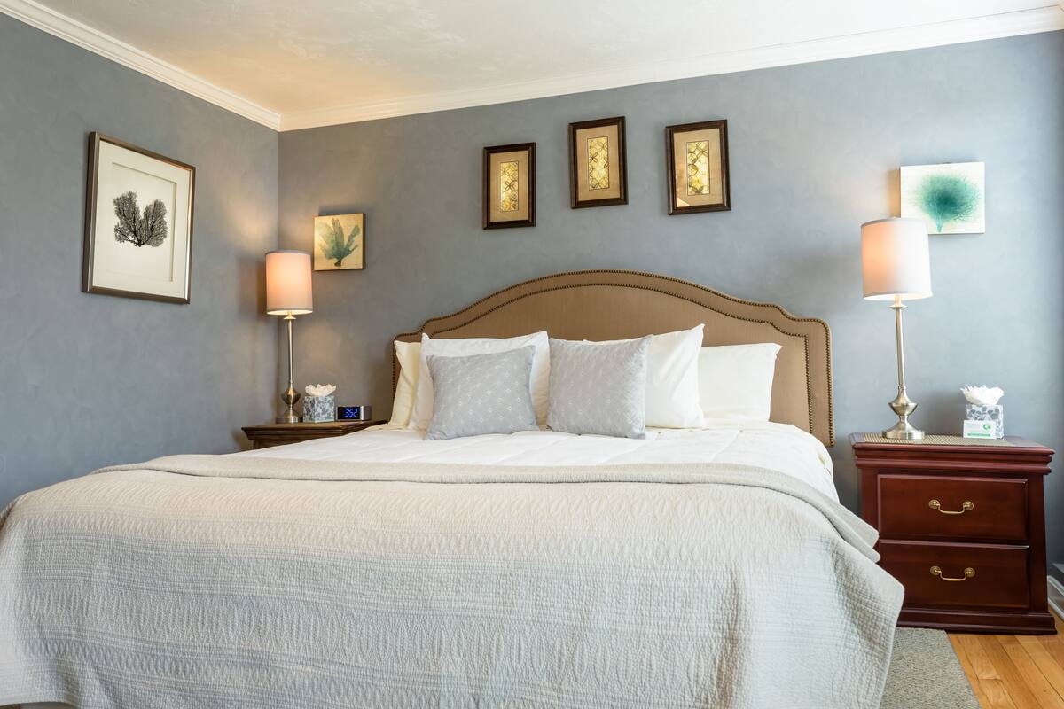 A neatly made bed with a white comforter and pillows, a wooden headboard, and a bedside table with a lamp and a clock.