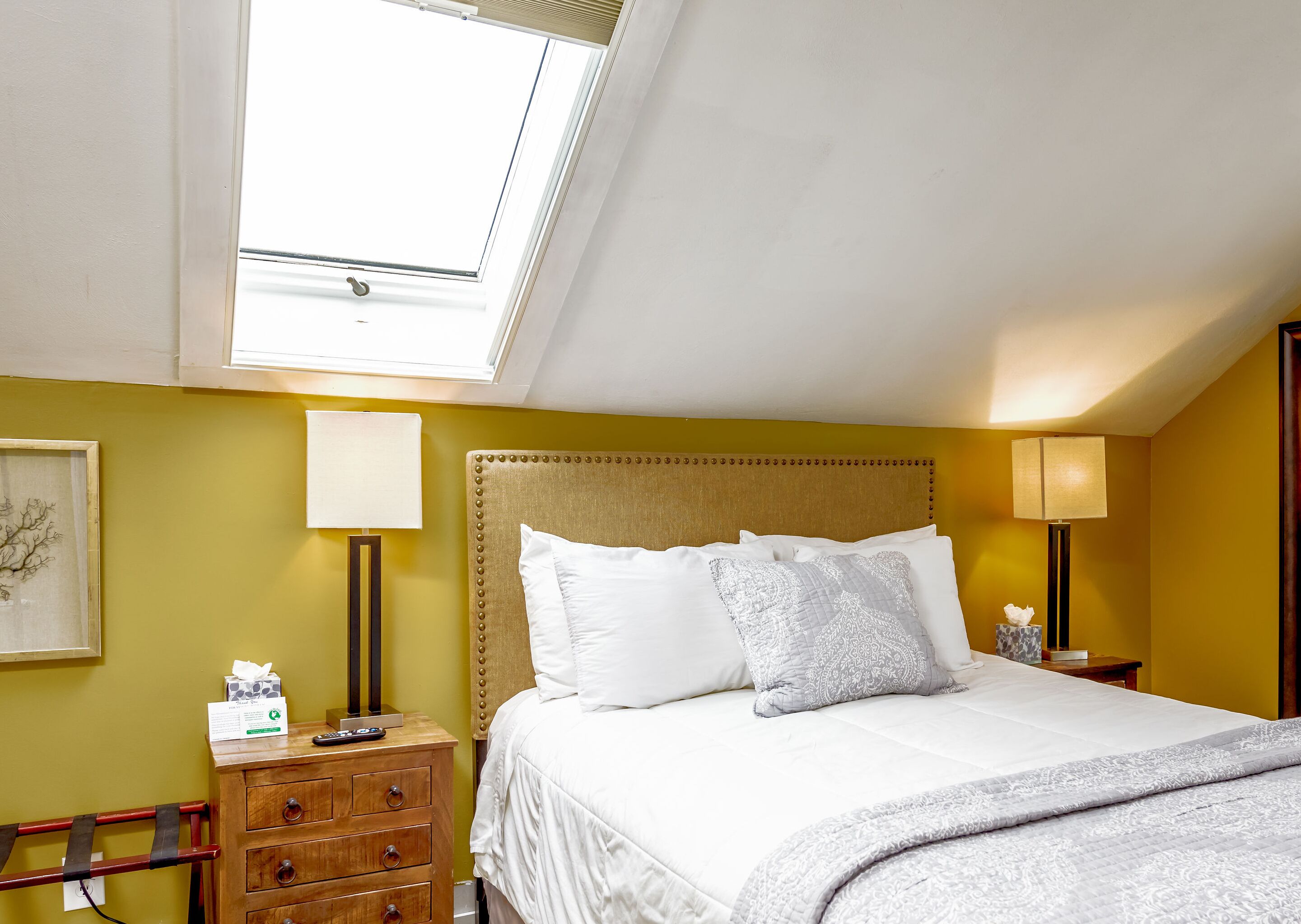 A hotel room with a bed, a nightstand, a lamp, a wall-mounted artwork, and a window with a view of the sky.