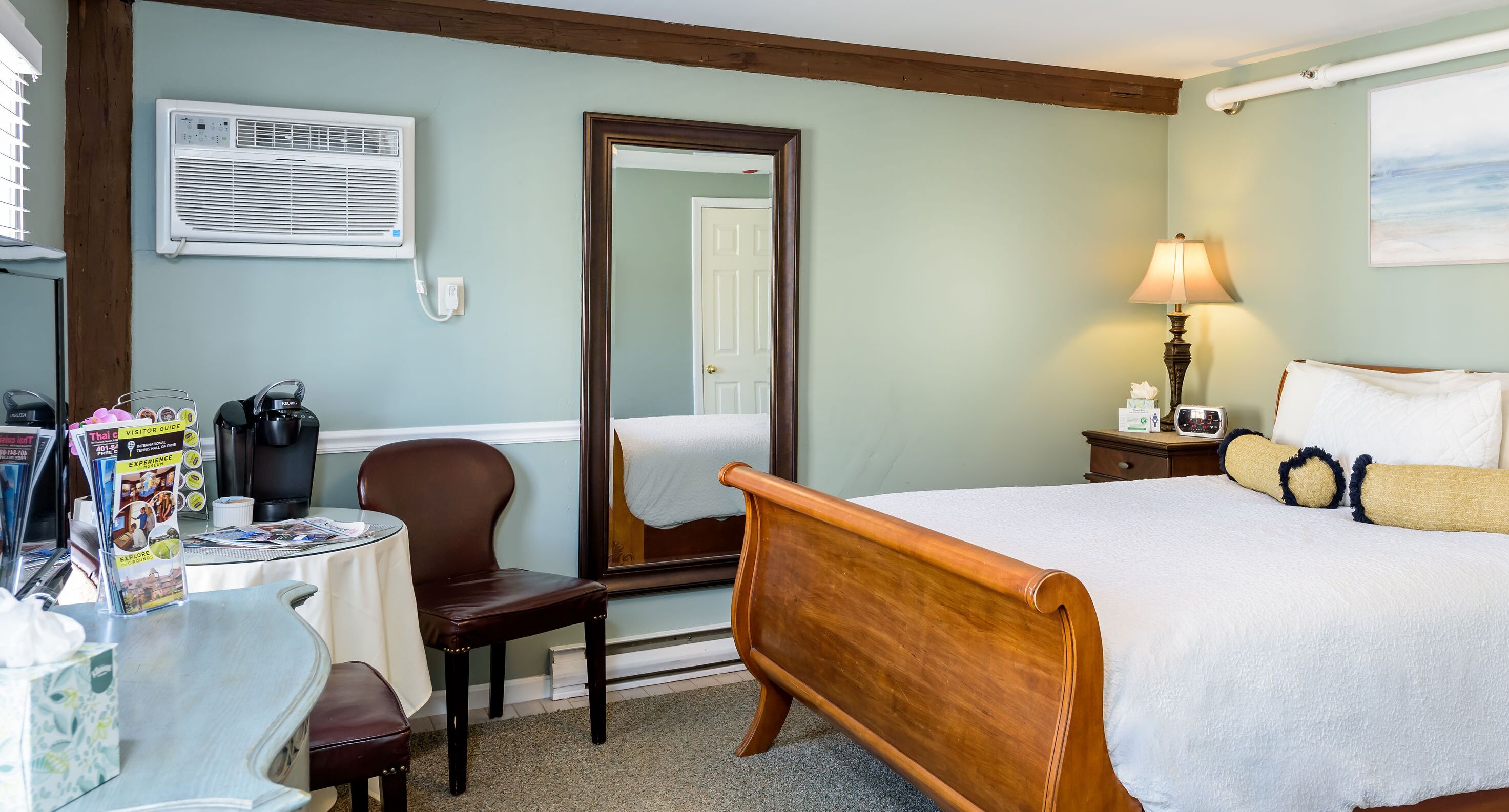 A hotel room with a bed, a chair, a table with brochures, a lamp, and a large mirror.