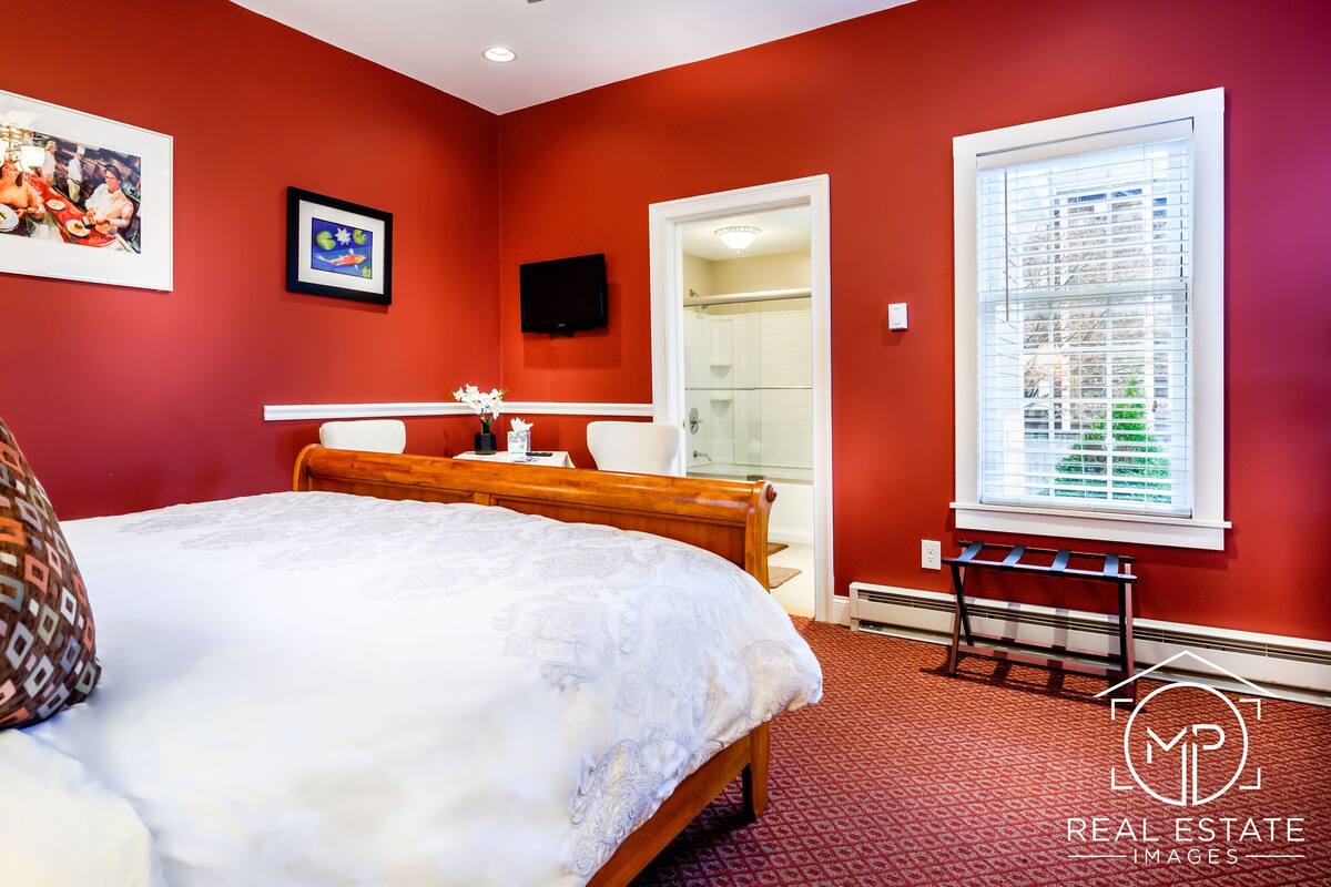 A bedroom with a bed, a TV, a window with blinds, and a bathroom visible through an open door.