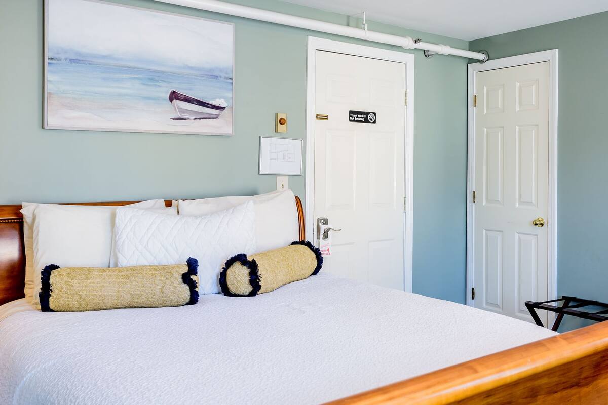 A neatly made bed with a wooden headboard, a framed painting of a boat on the wall, and a door with a no-smoking sign.