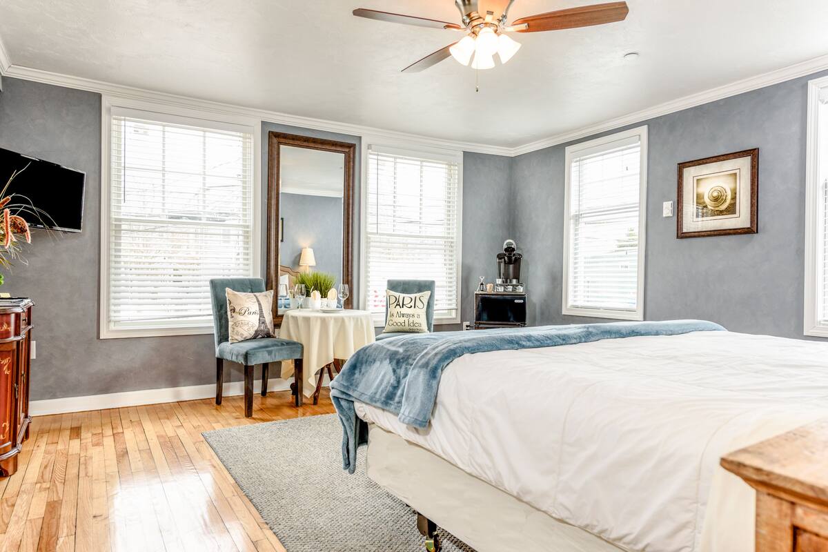 A bedroom with a large bed, a desk with a chair, a ceiling fan, and a TV mounted on the wall.