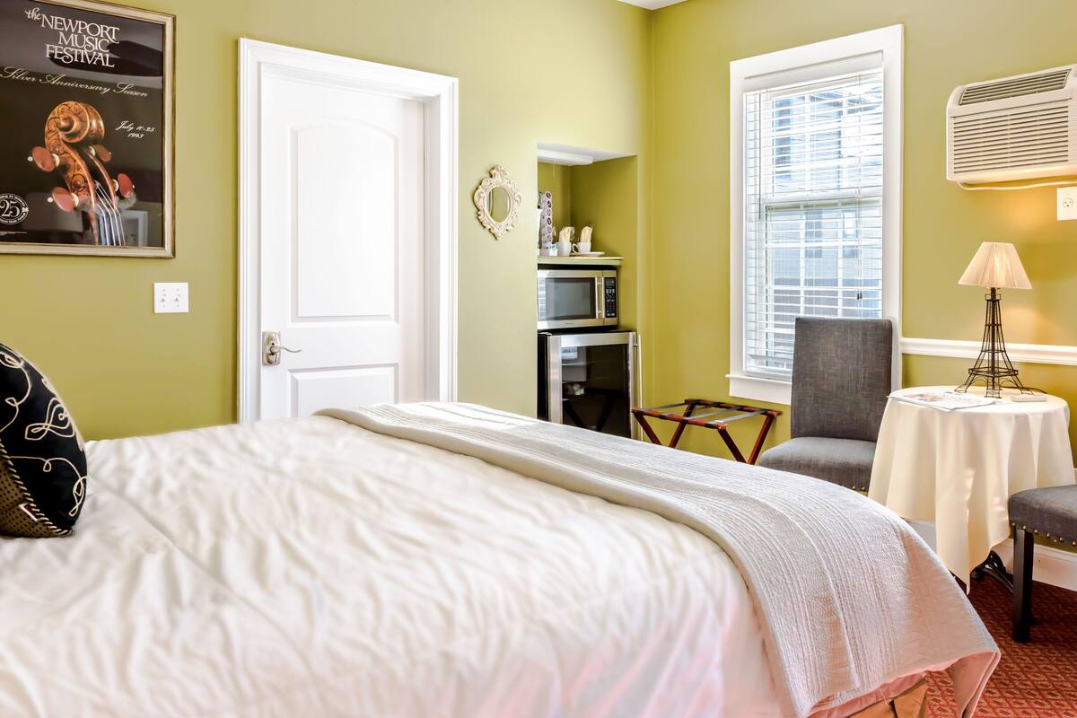 A hotel room with a bed, a chair, a table, a microwave, and a window with blinds.
