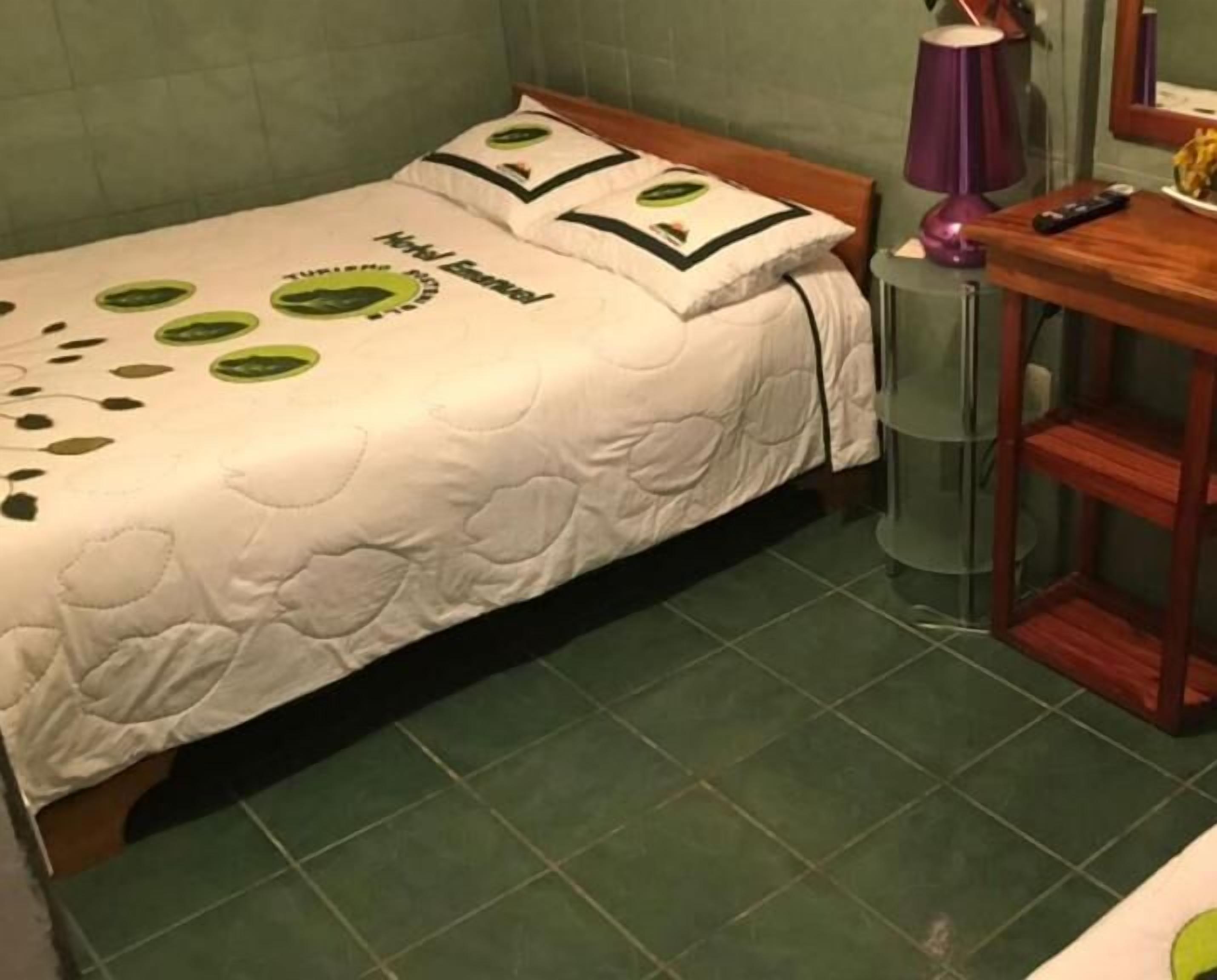 Un letto singolo con una coperta bianca e motivi a foglia verde, un comodino in legno con una lampada, e un tavolino con un telecomando.