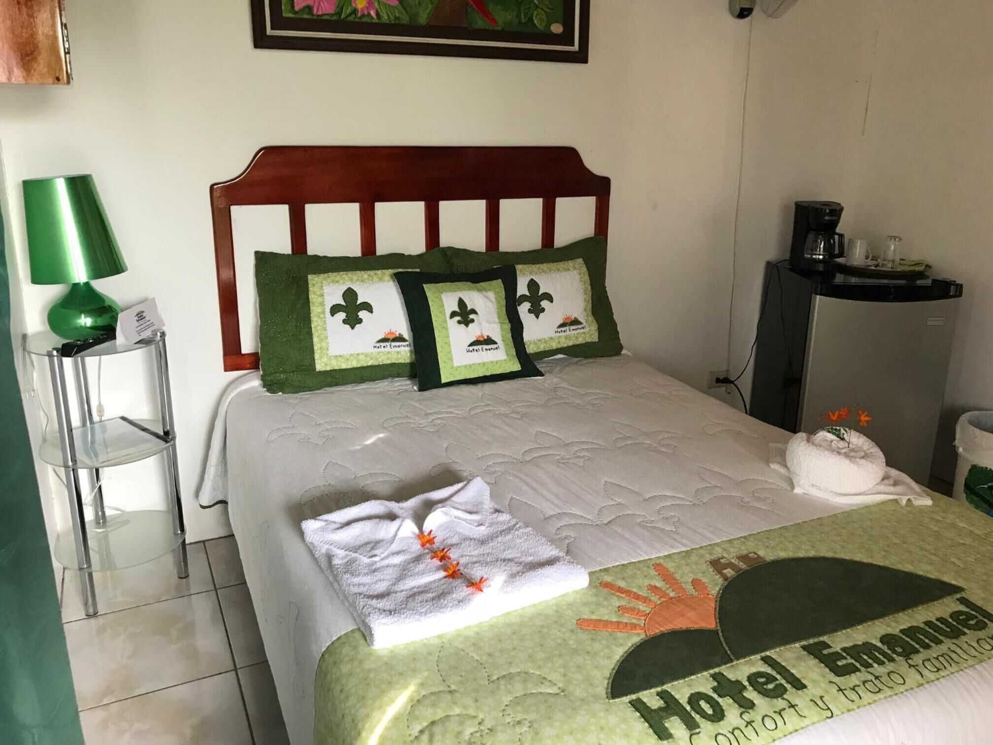 Una cama bien hecha con una manta verde, almohadas con diseño de fleur-de-lis y una toalla a un lado.