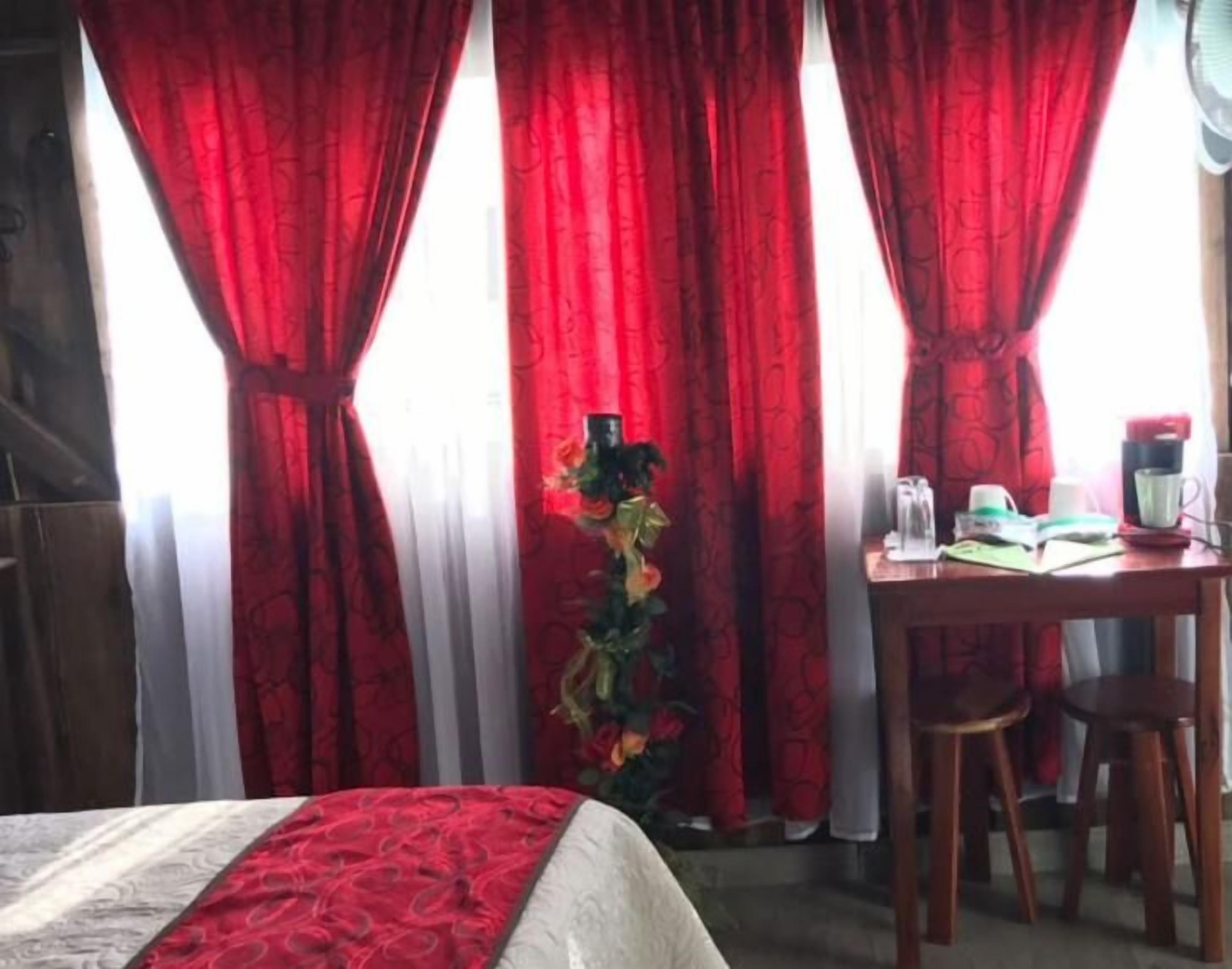 Een hotelkamer met een bed, een tafel met een kop en schotel, en rode gordijnen.