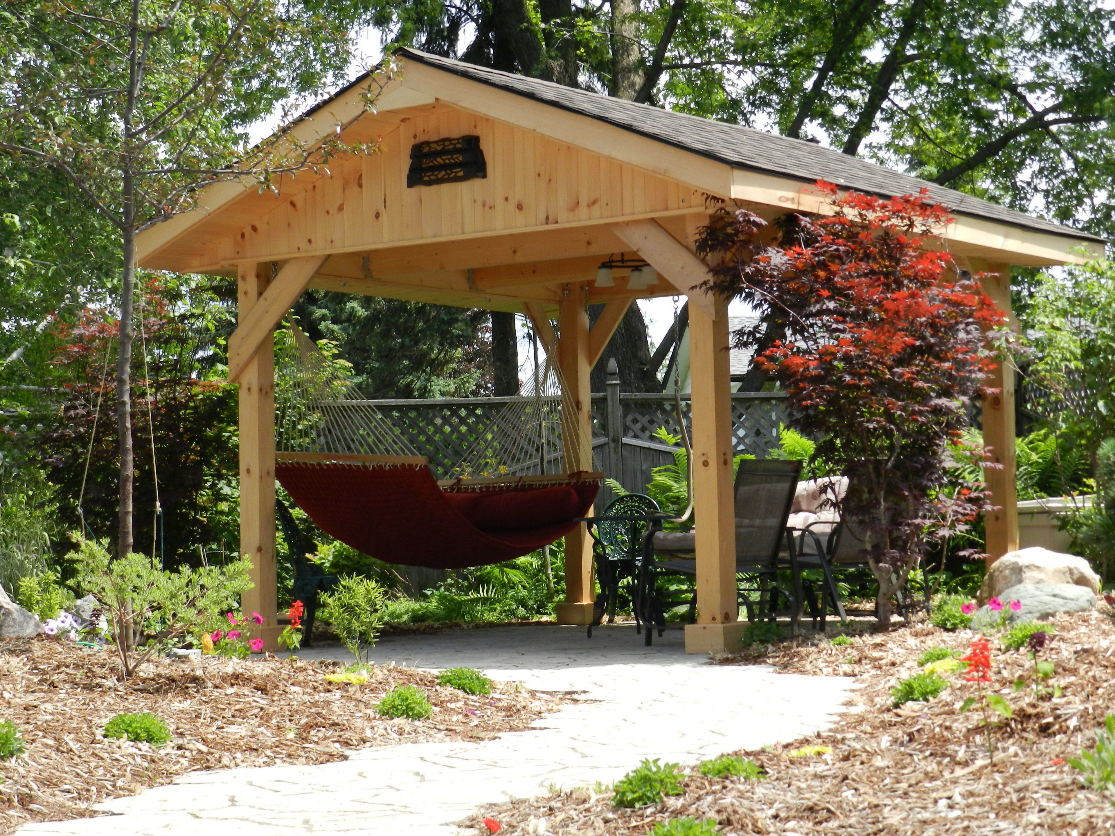 A wooden gazebo with a swing, surrounded by a garden with various plants and flowers.