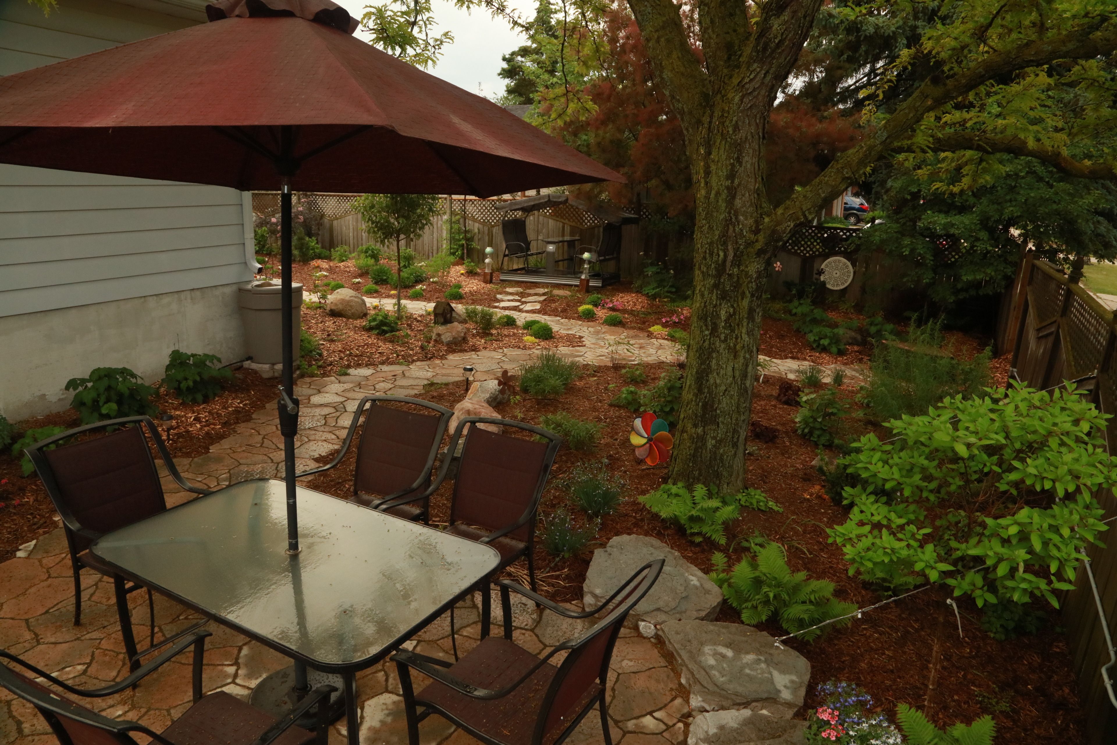 Um pátio externo com mesa e cadeiras, cercado por um jardim com diversas plantas e um cercado de madeira.