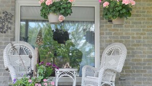 A porch with two white wicker rocking chairs, a potted plant with pink flowers, and a hanging flower basket. - A Garden Stroll B&B (Stratford)