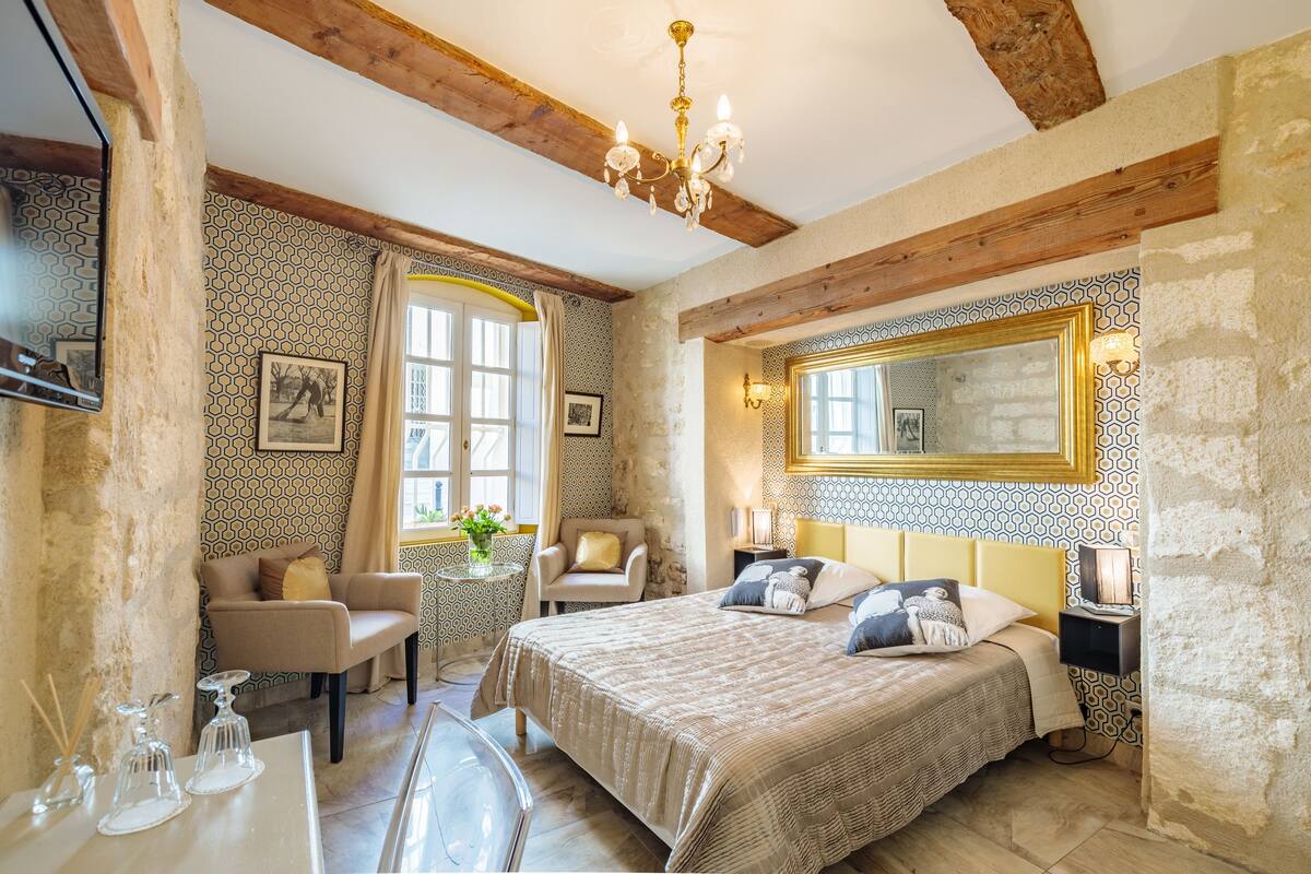 Une chambre avec un grand lit, un coin salon avec un fauteuil, une table avec des verres et un miroir au mur.