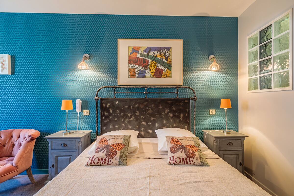 Une chambre avec un mur bleu à la texture particulière, un lit avec deux oreillers, une chaise, des tables de chevet avec des lampes et une œuvre d’art encadrée au-dessus du lit.