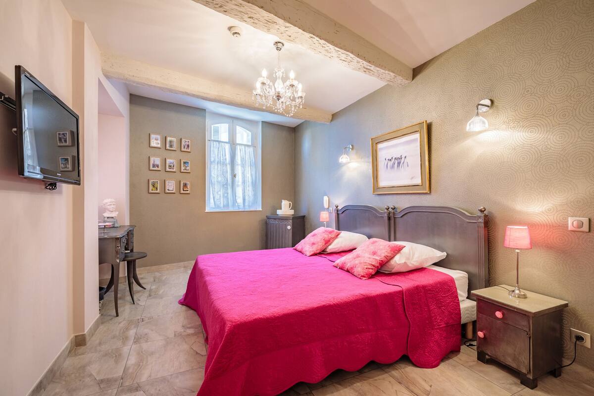 Une chambre à coucher comprenant un lit, une table de chevet, une commode, un téléviseur fixé au mur, un lustre, des œuvres d’art encadrées et une fenêtre avec des rideaux.