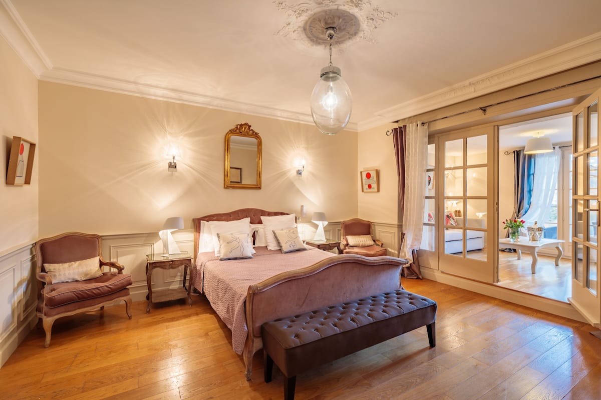 Une chambre spacieuse avec un grand lit, un banc, deux chaises, un miroir et une vue à travers une porte vitrée.