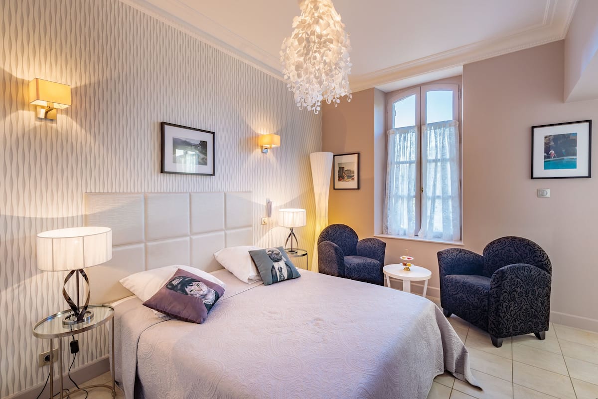 Une chambre d’hôtel avec un lit, deux chaises, une petite table et une fenêtre avec des rideaux.