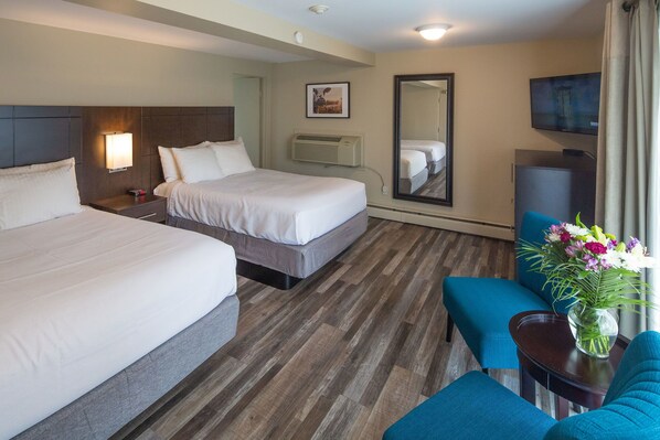 A hotel room with two beds, a TV, a mirror, a desk with a vase of flowers, and a blue chair.
