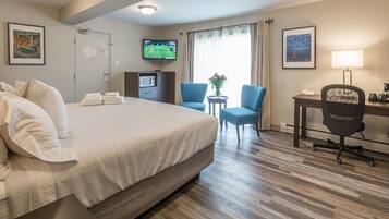 Quarto de hotel com uma cama grande, uma escrivaninha com cadeira, uma televisão e uma pequena mesa com abajur.