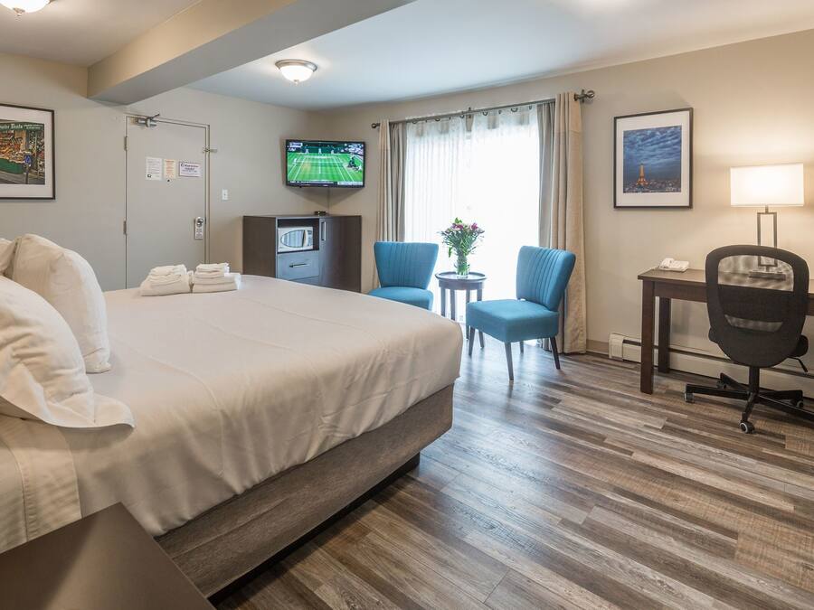 Quarto de hotel com uma cama grande, uma escrivaninha com cadeira, uma televisão e uma pequena mesa com abajur.