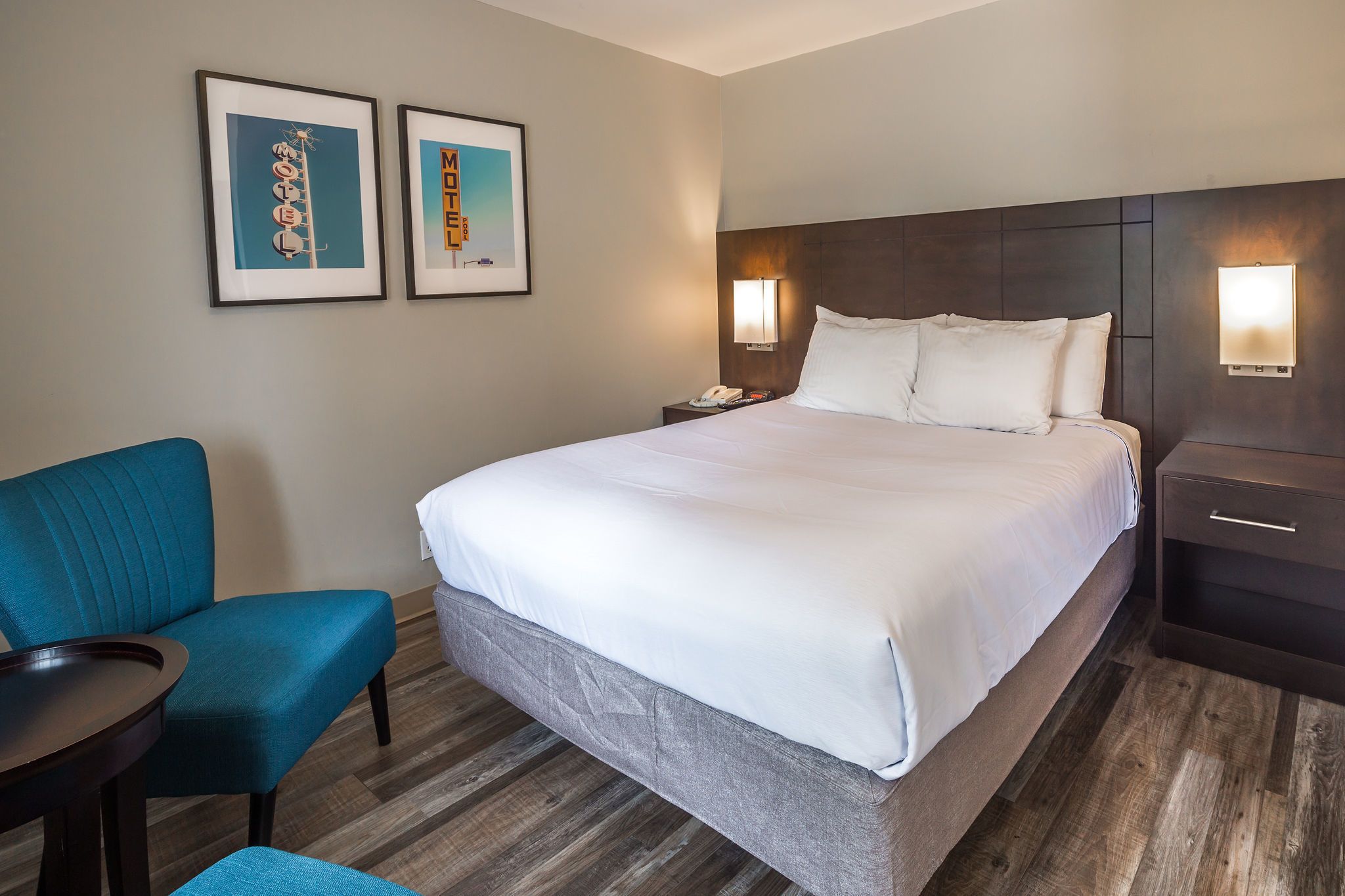 A hotel room with a large bed, a blue armchair, a small table, and two framed pictures on the wall.