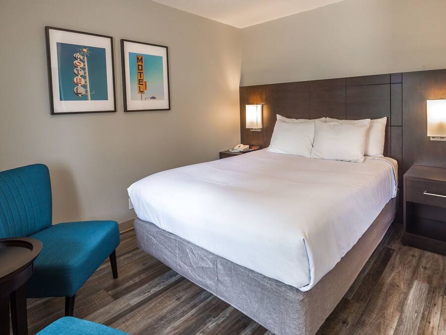 A hotel room with a large bed, a blue armchair, a small table, and two framed pictures on the wall.