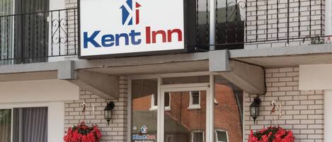 Letrero de Kent Inn sobre la entrada de un edificio con un balcón, dos cestas de flores y un letrero que indica los números de habitación de 107 a 117, no se admiten mascotas y no se permite fumar.
