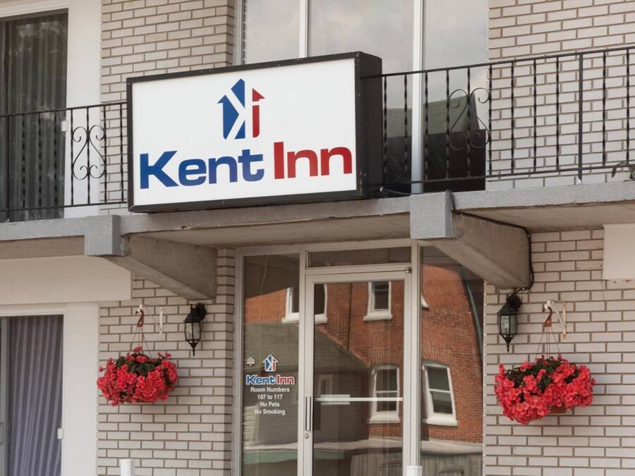 Une enseigne indiquant « Kent Inn » est placée au-dessus de l’entrée d’un immeuble, qui est doté d’un balcon, de deux jardinières fleuries et d’un panneau indiquant les numéros de chambre, du 107 au 117, ainsi que les mentions « animaux non admis » et « il est interdit de fumer ».