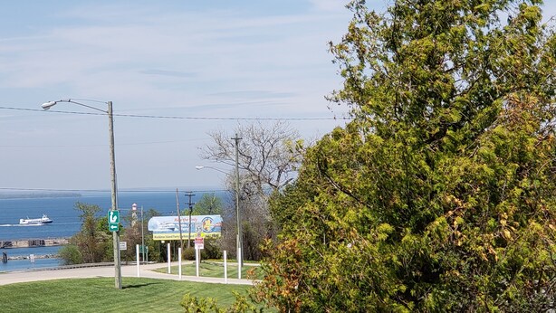 A grassy area with a sign for Mackinac Island Ferry, a body of water, and a distant view of a lighthouse. - Aurora Borealis Motel (St Ignace)