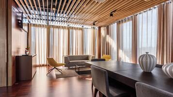 Una habitación de hotel moderna con techo de madera, un sofá, una mesa de comedor y un gran ventanal con cortinas.