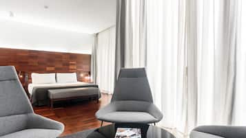 Une chambre d’hôtel moderne avec une tête de lit en bois, un grand lit, un fauteuil gris, une petite table avec un magazine et de grandes fenêtres ornées de rideaux légers.