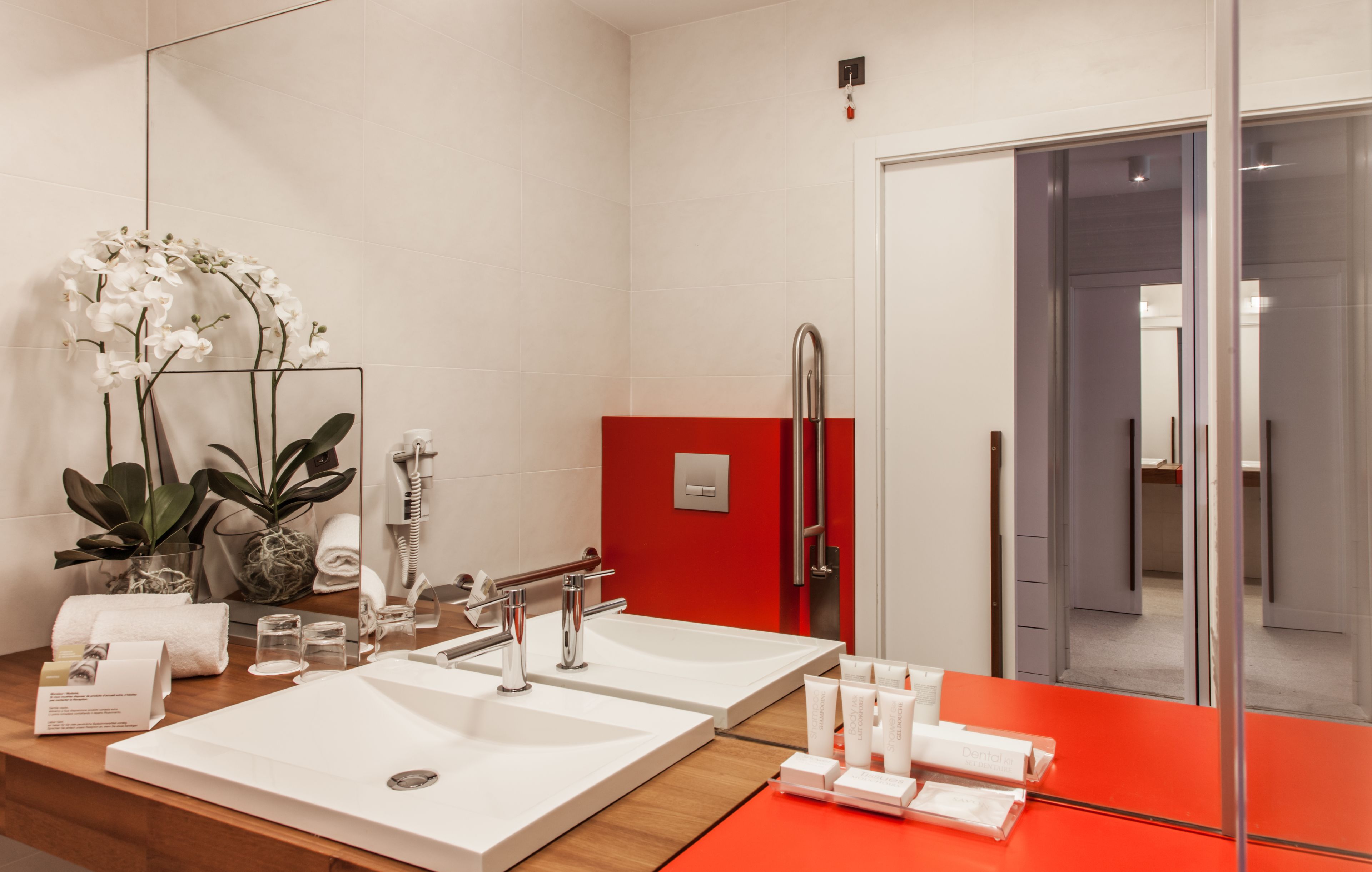 Un baño moderno con dos lavamanos, un espejo, una pared de acento roja y una cabina de ducha con mampara de vidrio.