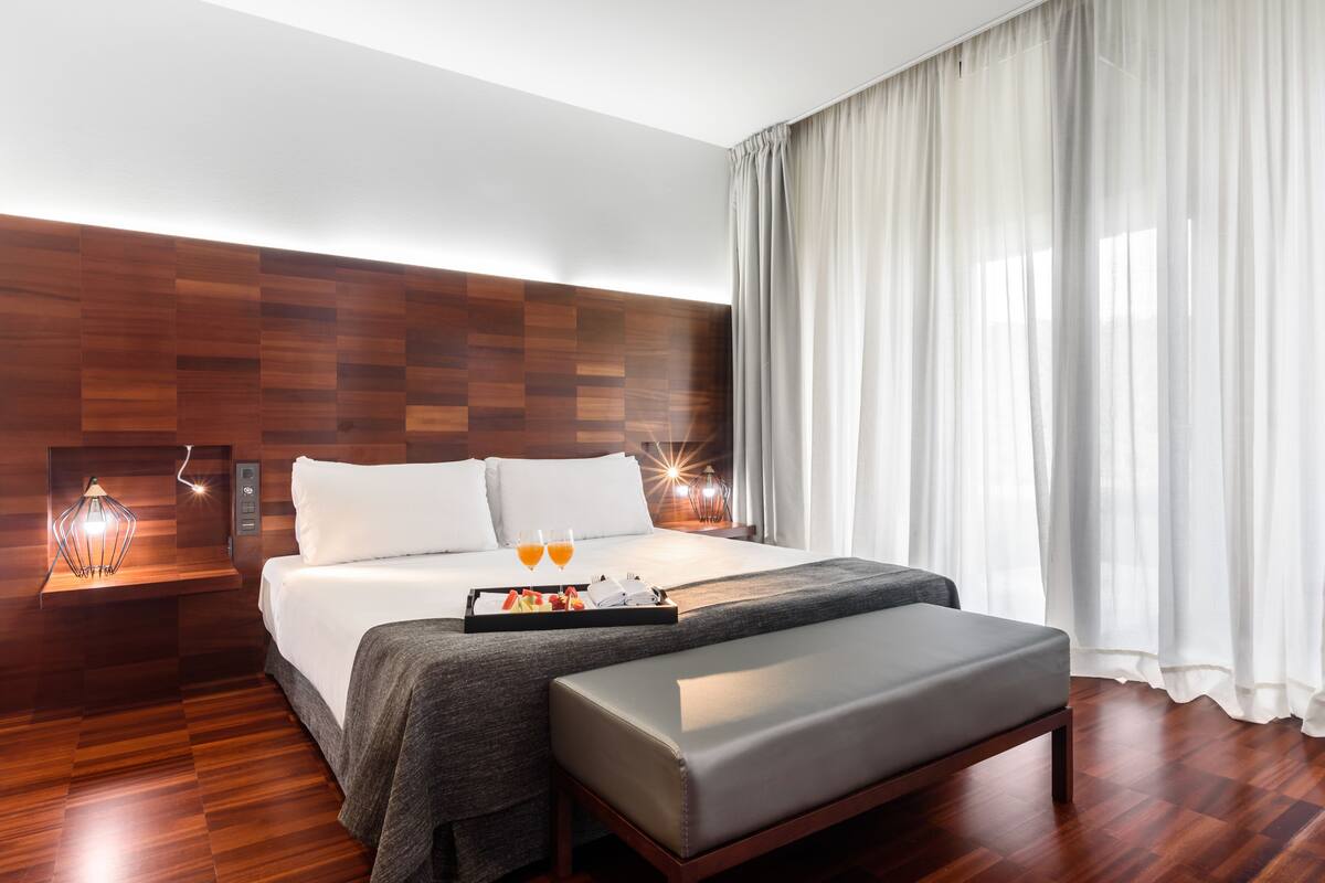 Quarto de hotel moderno com cabeceira de madeira, uma cama grande com colchão cinza, mesa de cabeceira com abajur e bandeja com frutas e copos de suco de laranja.