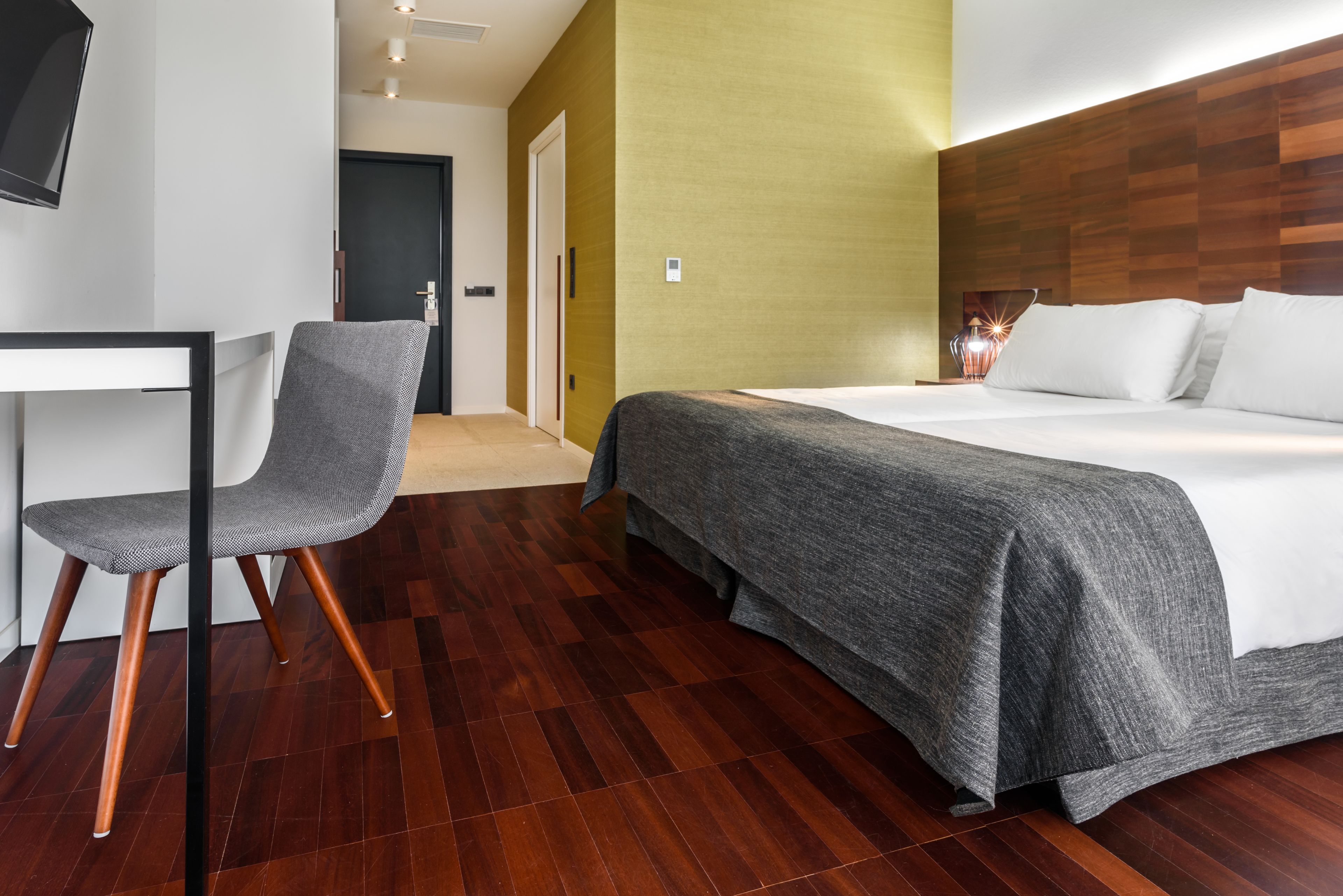 Una habitació d'hotel moderna amb terra de fusta, una cadira tapissada de color gris, un llit amb roba blanca i un panell de fusta al capçal.