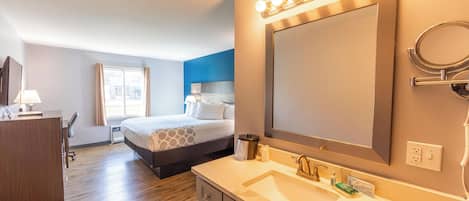 A hotel room with a large bed, a bedside table, a mirror, a vanity with a sink, and a window with curtains.