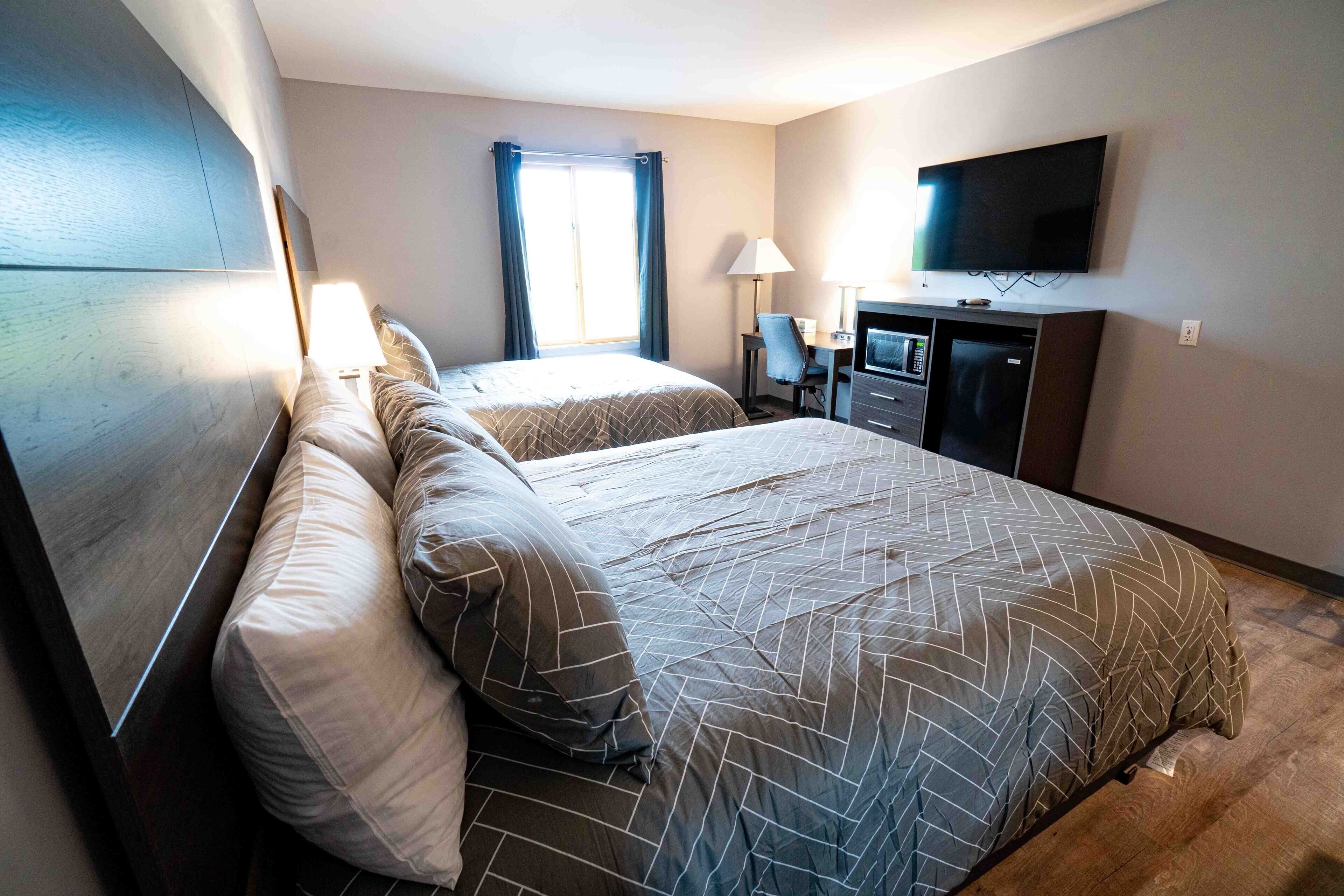 Ein Hotelzimmer mit zwei Betten, einem Schreibtisch mit Stuhl, einem Fernseher und einem großen Fenster mit Vorhängen.
