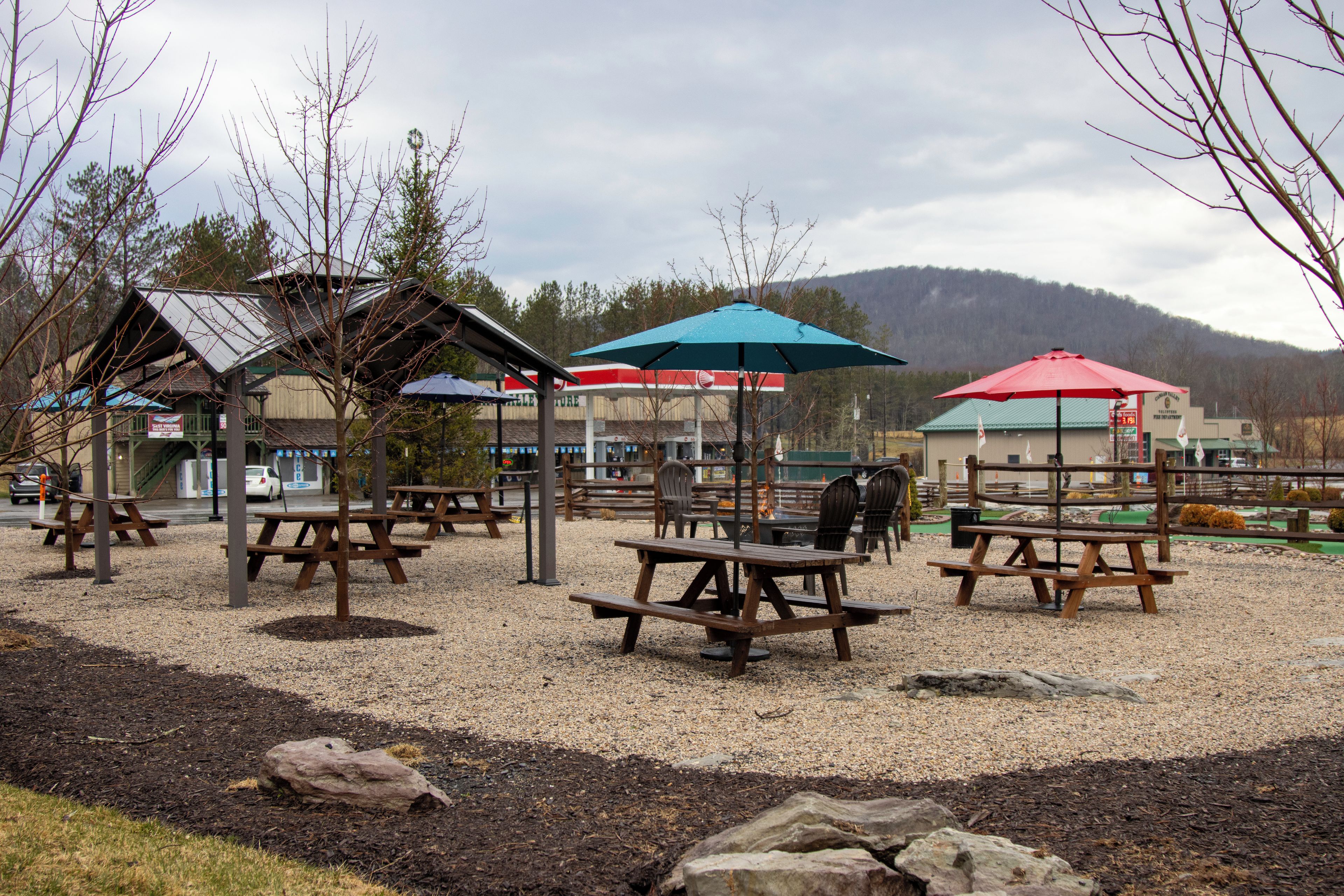 An outdoor seating area with picnic tables and umbrellas, surrounded by trees and a mountain in the background.