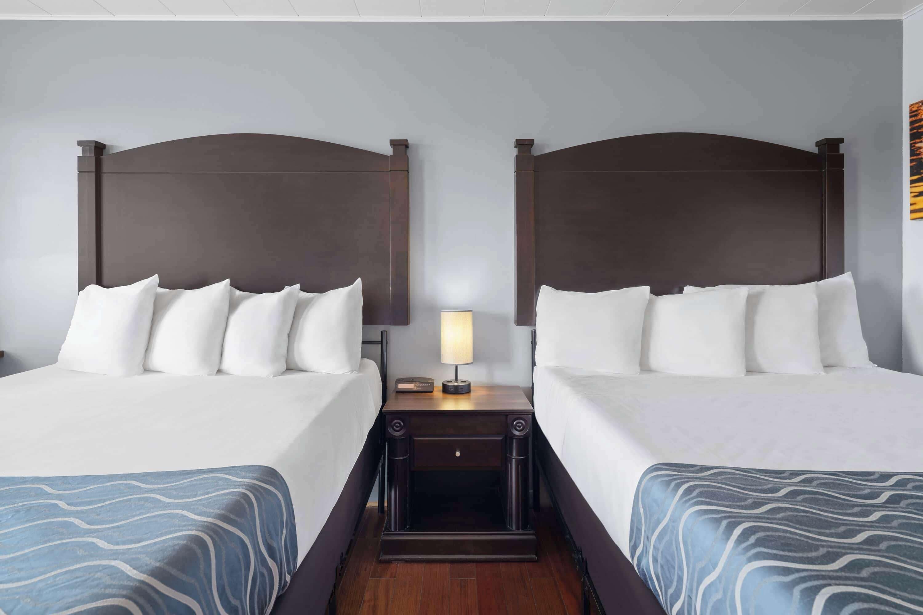 A hotel room with two beds, a nightstand with a lamp, and a wooden floor.