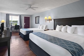 A hotel room with two beds, a desk with a computer, a chair, a window with curtains, a door, and a ceiling fan.